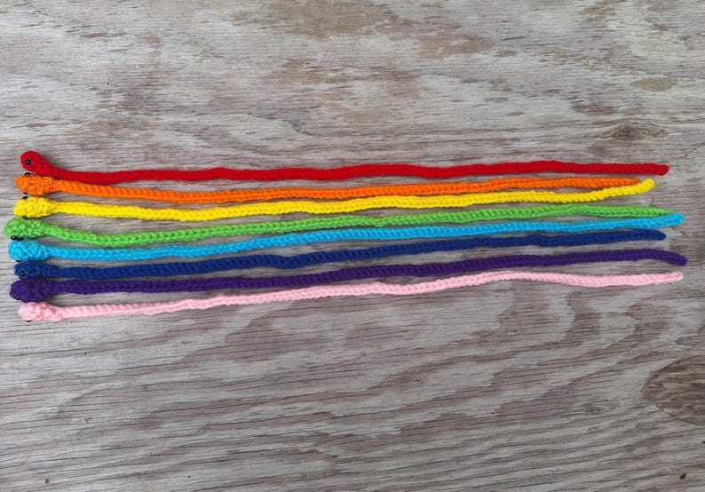 May include: A set of ten crocheted rainbow-colored snakes with black eyes. The snakes are arranged in a row from red to pink, with each snake slightly shorter than the one before it.