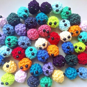 May include: A collection of small, round, crocheted yarn spheres in a variety of vibrant colors including red, blue, green, yellow, purple, and pink. Each sphere features two small, shiny black plastic beads attached to the front as eyes.