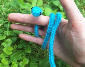 Könnte beinhalten: Eine gehäkelte blaue Schlange mit schwarzen Augen. Die Schlange ist um einen Finger gewickelt.