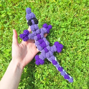 May include: A handmade crochet lizard in shades of purple, black, and white, with a textured, three-dimensional design. The lizard is held against a backdrop of green grass, showcasing its intricate details and craftsmanship.