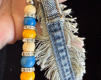 May include: A handmade keychain featuring a denim fabric accent with frayed edges and a beaded charm. The charm includes silver-toned beads, and colorful wooden beads in yellow, blue, and cream. A silver keyring completes the design.
