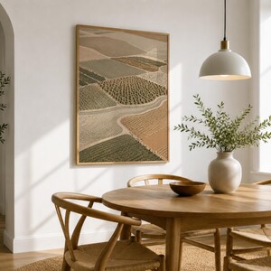 Pode incluir: Uma impress&atilde;o de arte de parede vertical emoldurada com uma vista a&eacute;rea de campos agr&iacute;colas em tons de bege, verde-oliva e castanho. A obra exibe um mosaico de linhas de cultivo texturizadas e caminhos sinuosos, numa moldura de madeira clara.