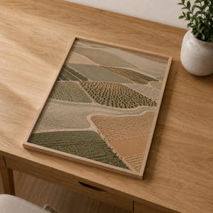 Pode incluir: Uma impress&atilde;o retangular emoldurada com uma vista a&eacute;rea de campos agr&iacute;colas em tons de verde s&aacute;lvia, oliva, bege e ocre. A obra exibe um mosaico de padr&otilde;es texturizados que representam v&aacute;rias culturas dentro de uma moldura de madeira clara.