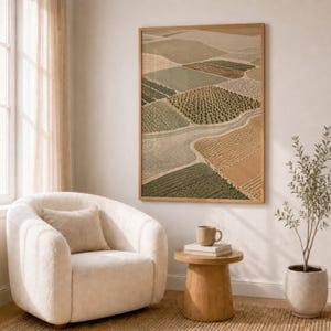 Pode incluir: Uma impress&atilde;o de arte de parede emoldurada com uma vista a&eacute;rea de campos agr&iacute;colas em tons de verde, bege e castanho. A obra de arte est&aacute; exposta numa parede acima de uma poltrona creme, uma pequena mesa de apoio em madeira com uma caneca e uma planta em vaso.