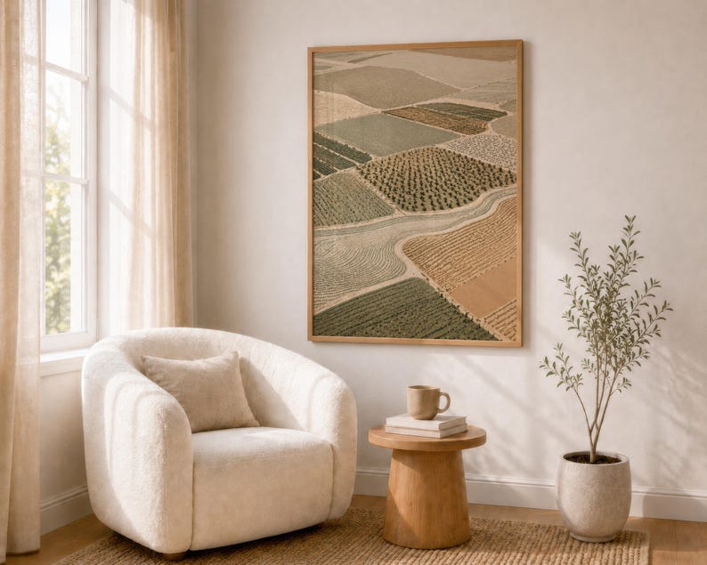 Pode incluir: Uma impress&atilde;o de arte de parede emoldurada com uma vista a&eacute;rea de campos agr&iacute;colas em tons de verde, bege e castanho. A obra de arte est&aacute; exposta numa parede acima de uma poltrona creme, uma pequena mesa de apoio em madeira com uma caneca e uma planta em vaso.