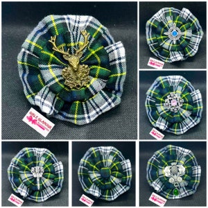 May include: A selection of tartan fabric brooches in green, blue, yellow, and white. Each brooch has a different decorative centrepiece, including a stag, a blue gem, a pink gem, and a silver design with a green gem. The brooches are from Girls Glamour Accessories.