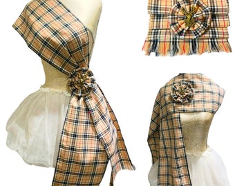 May include: A plaid sash with a gold and red pin, tied in a bow. The sash is beige, black, and red with a plaid pattern. The sash is displayed on a white mannequin.