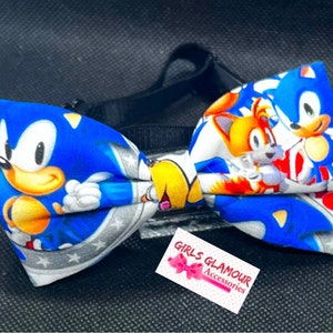 May include: A bow tie with a blue Sonic the Hedgehog and Tails design. The bow tie has a black adjustable strap and a white tag with the text "GIRLS GLAMOUR ACCESSORIES". The bow tie is made of fabric.