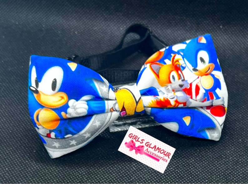 May include: A blue bow tie featuring Sonic the Hedgehog and Tails characters. The bow tie has a black adjustable strap and a white tag with the text "GIRLS GLAMOUR ACCESSORIES". The bow tie is made of fabric.