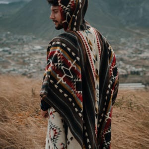 May include: A person wearing a hooded poncho with a colorful geometric pattern. The poncho is brown, white, red, and yellow. The person is standing in a field of tall grass.