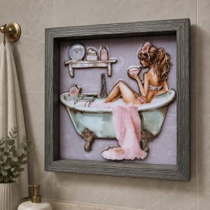 May include: A framed wall art piece depicts a woman relaxing in a vintage bathtub. The artwork features a light blue tub with ornate feet, a pink towel, and a shelf with accessories. The frame is a weathered gray.
