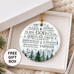 May include: A round white ceramic ornament featuring a mountain and pine tree landscape with printed text including ABBA, EL ELYON, ADONAI, EL ROI, EL OLAM, JEHOVAH JIREH, JEHOVAH RAAH, EL SHADDAI, JEHOVAH NISSI, and YAHWEH, suspended by a gold string in a white box.