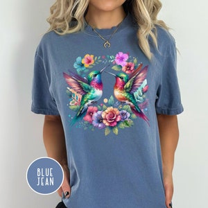 May include: A blue jean t-shirt with a colorful floral design featuring two hummingbirds facing each other. The hummingbirds are surrounded by pink, purple, and yellow flowers.