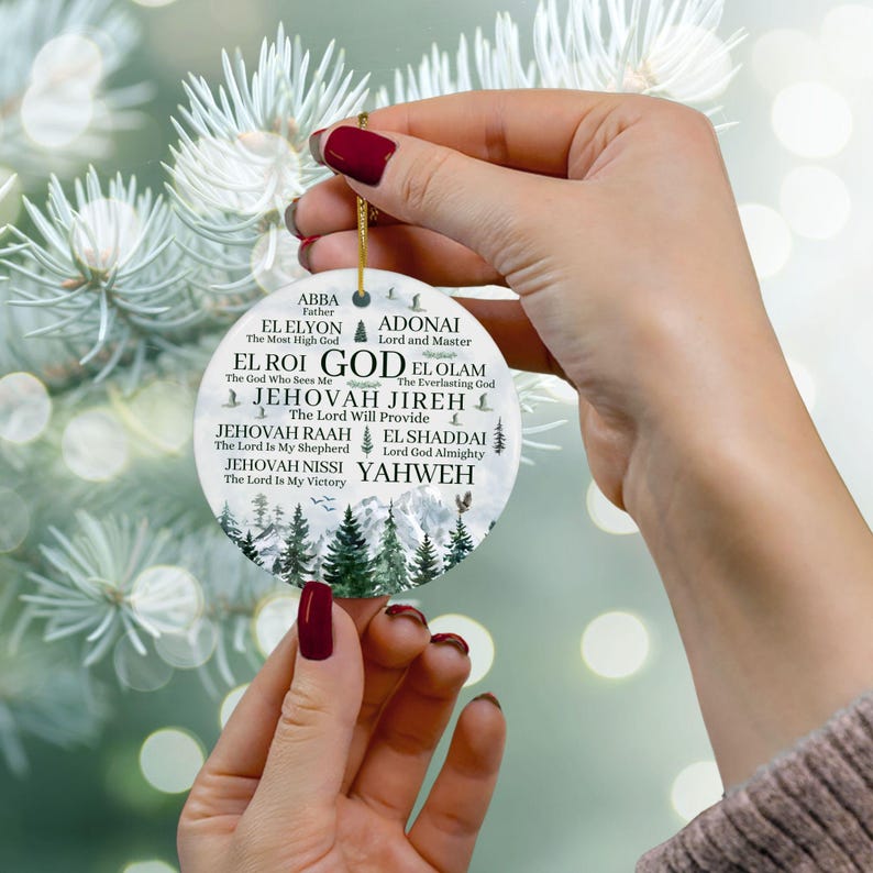 May include: A round white ceramic ornament featuring a mountain and pine tree landscape at the bottom. The text includes names of God such as ABBA, EL ELYON, ADONAI, EL ROI, GOD, EL OLAM, JEHOVAH JIREH, JEHOVAH RAAH, EL SHADDAI, JEHOVAH NISSI, and YAHWEH.