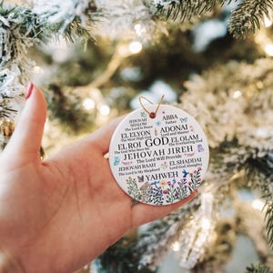 May include: A round white ceramic ornament featuring a gold string loop. The surface displays various names of God in black text, including Jehovah Shalom, El Elyon, El Roi, Jehovah Jireh, Jehovah Raah, Abba, Adonai, El Olam, El Shaddai, and Yahweh.