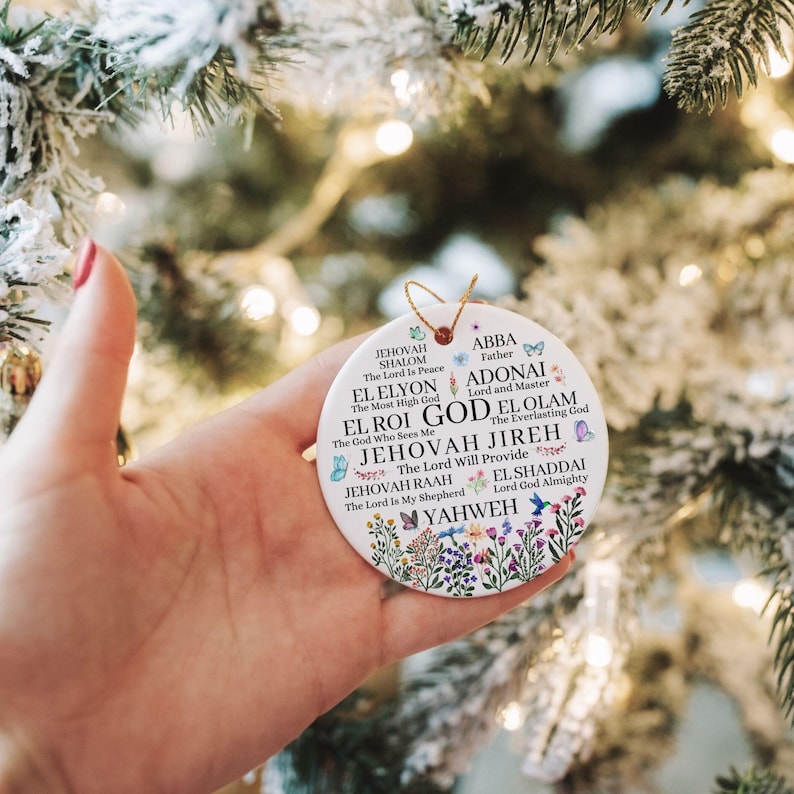 May include: A round white ceramic ornament featuring a gold string loop. The surface displays various names of God in black text, including Jehovah Shalom, El Elyon, El Roi, Jehovah Jireh, Jehovah Raah, Abba, Adonai, El Olam, El Shaddai, and Yahweh.