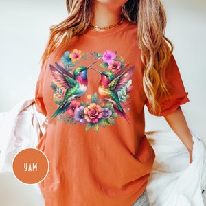May include: A burnt orange t-shirt with a colorful hummingbird design. The hummingbirds are facing each other and are surrounded by flowers.