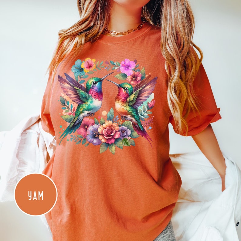 May include: A burnt orange t-shirt with a colorful hummingbird design. The hummingbirds are facing each other and are surrounded by flowers.