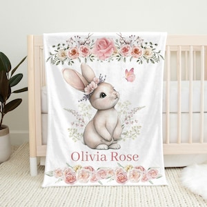Peut inclure: Une couverture de b&eacute;b&eacute; blanche avec une illustration &agrave; l'aquarelle d'un lapin portant une couronne de fleurs, un papillon rose et des motifs floraux. Le nom "Olivia Rose" est imprim&eacute; en script rose. La couverture est expos&eacute;e sur un berceau.