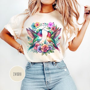 May include: A white t-shirt with a colorful hummingbird design. The hummingbirds are surrounded by flowers and leaves. The shirt is available in ivory.