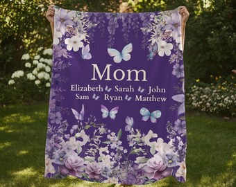 May include: A purple blanket with floral and butterfly designs. The word "Mom" is centered, with names below. The blanket has a watercolor style with white, purple, and blue flowers and butterflies. The blanket is held up in a grassy outdoor setting.