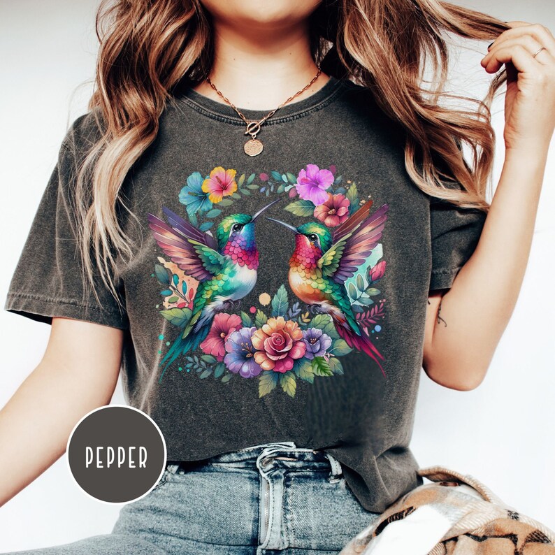 May include: A dark gray t-shirt with a colorful floral design featuring two hummingbirds. The hummingbirds are facing each other and have iridescent green, blue, and red feathers. The flowers are pink, purple, and yellow.