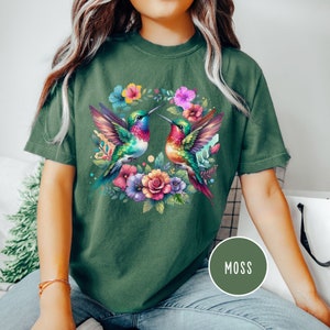May include: A green t-shirt with a colorful floral design featuring two hummingbirds. The hummingbirds are facing each other and have their beaks touching. The flowers are in shades of pink, purple, yellow, and green. The text "MOSS" is printed in white on a green circle in the lower right corner of the image.