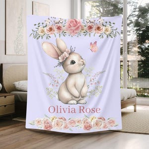 Peut inclure: Une couverture lilas avec une illustration &agrave; l'aquarelle d'un lapin portant une couronne de fleurs, un papillon et des bordures florales. Le nom "Olivia Rose" est &eacute;crit en rose. La couverture est pos&eacute;e sur un lit.