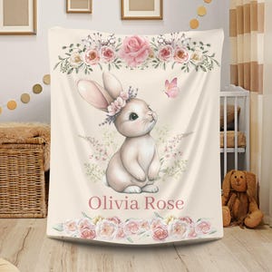 Peut inclure: Une couverture pour b&eacute;b&eacute; cr&egrave;me avec une illustration &agrave; l'aquarelle d'un lapin portant une couronne florale. La couverture est orn&eacute;e de roses roses et d'un papillon. Le nom "Olivia Rose" est imprim&eacute; en rose.
