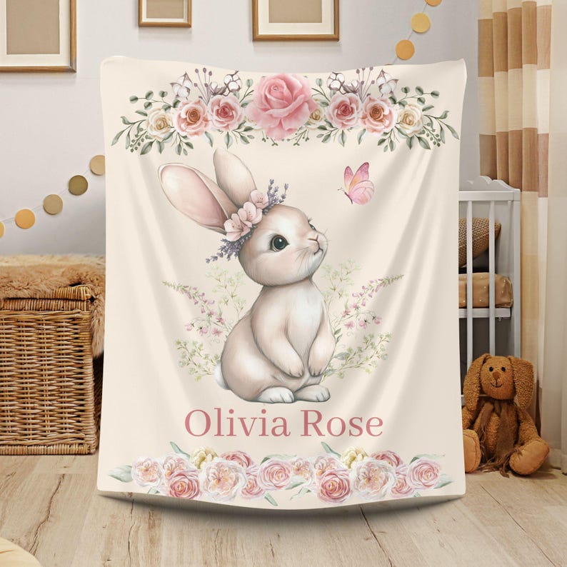 Peut inclure: Une couverture pour b&eacute;b&eacute; cr&egrave;me avec une illustration &agrave; l'aquarelle d'un lapin portant une couronne florale. La couverture est orn&eacute;e de roses roses et d'un papillon. Le nom "Olivia Rose" est imprim&eacute; en rose.