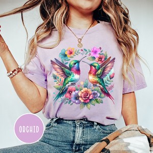 May include: A purple t-shirt with a floral design featuring two hummingbirds with colorful wings and beaks. The hummingbirds are surrounded by pink, yellow, and orange flowers.
