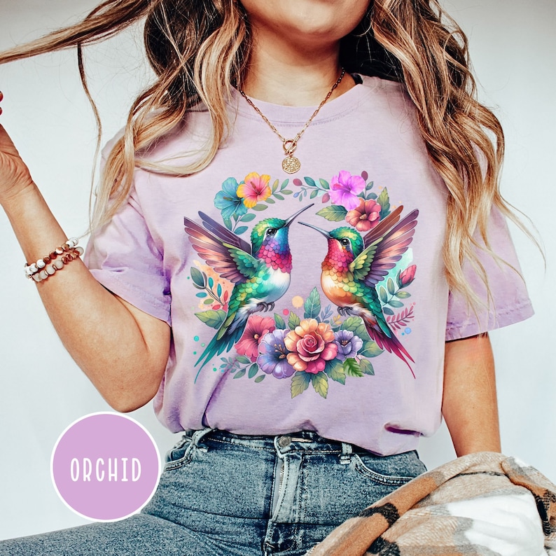 May include: A purple t-shirt with a floral design featuring two hummingbirds with colorful wings and beaks. The hummingbirds are surrounded by pink, yellow, and orange flowers.