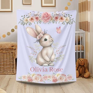 Peut inclure: Une couverture de b&eacute;b&eacute; violet clair avec une illustration &agrave; l'aquarelle d'un lapin portant une couronne de fleurs. Le motif comprend des roses roses, un papillon et le nom "Olivia Rose" en &eacute;criture rose. La couverture a des bordures florales.