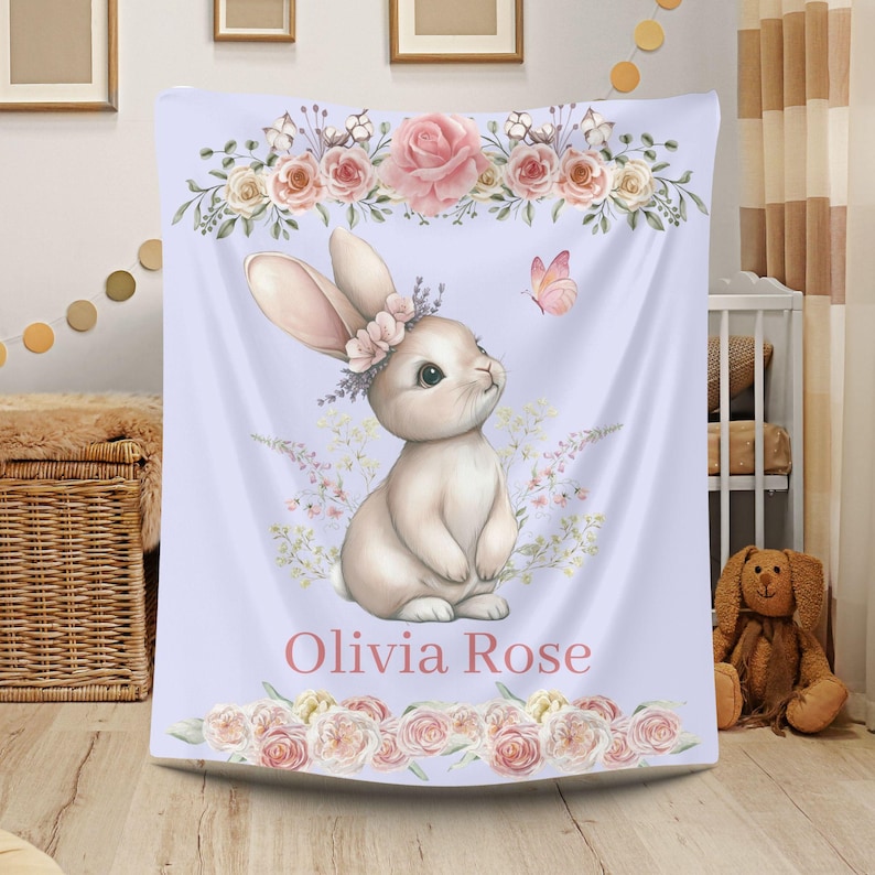 Peut inclure: Une couverture de b&eacute;b&eacute; violet clair avec une illustration &agrave; l'aquarelle d'un lapin portant une couronne de fleurs. Le motif comprend des roses roses, un papillon et le nom "Olivia Rose" en &eacute;criture rose. La couverture a des bordures florales.