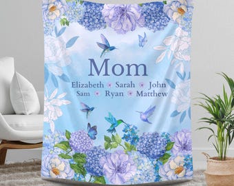 May include: A light blue blanket with a floral design featuring hydrangeas and peonies. The word "Mom" is centered, with names below. Hummingbirds and butterflies are scattered across the blanket. The blanket is displayed in a living room setting.