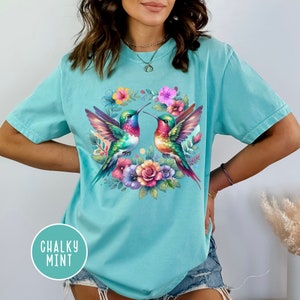 May include: A turquoise blue t-shirt with a colorful floral wreath design featuring two hummingbirds. The hummingbirds are facing each other and have iridescent feathers. The wreath is made up of pink, purple, and yellow flowers.