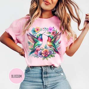 May include: A pink t-shirt with a colorful floral design featuring two hummingbirds facing each other. The design includes a wreath of flowers and leaves.