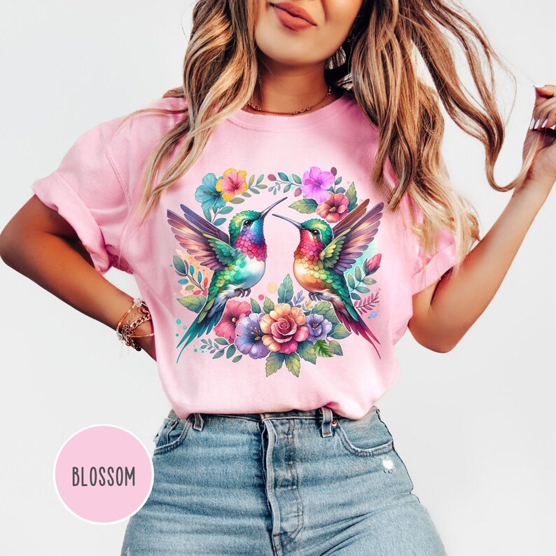 May include: A pink t-shirt with a colorful floral design featuring two hummingbirds facing each other. The design includes a wreath of flowers and leaves.