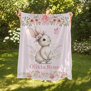 Peut inclure: Une couverture rose clair pour b&eacute;b&eacute; avec une illustration &agrave; l'aquarelle d'un lapin portant une couronne florale. La couverture est orn&eacute;e de roses et du nom "Olivia Rose". Un papillon est &eacute;galement pr&eacute;sent.