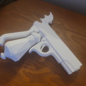 Op de afbeelding: Een witte, 3D-geprinte speelgoedpistool met een uniek ontwerp. Het speelgoedpistool heeft een trekker, handvat en een decoratief element aan de bovenkant. Het object is gemaakt van een solide, ondoorzichtig materiaal en ligt op een houten oppervlak.