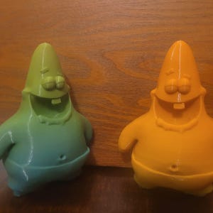 Op de afbeelding: Twee 3D-geprinte figuren van Patrick Star uit Spongebob Squarepants. Eén is een verloop van groen naar blauw, de andere is oranje. Beide hebben een lachend gezicht, armen en een navel. Ze staan op een houten oppervlak.