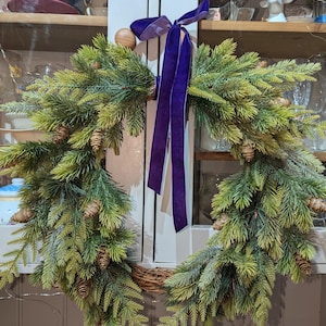 May include: A Christmas wreath featuring artificial evergreen foliage and pine cones. It is decorated with a purple velvet ribbon and a wooden ball. The wreath is displayed on a white cabinet.