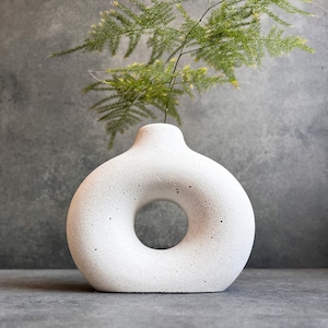 May include: A white, donut-shaped vase with a textured surface. The vase has a circular opening in the center and a narrow neck. Green fern-like foliage is placed in the vase, creating a minimalist floral arrangement. The vase is a decorative home accent.