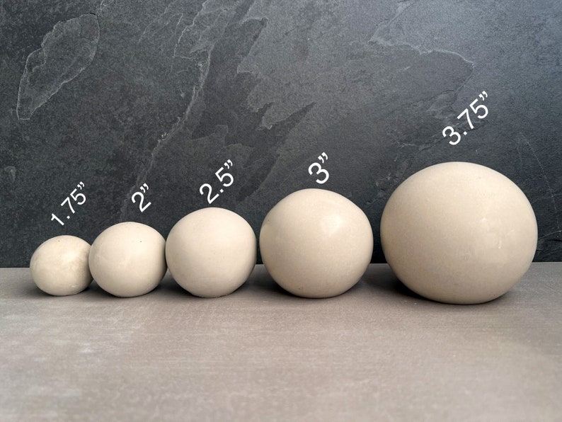 Puede incluir: Cinco esferas blanquecinas de varios tama&ntilde;os, que van desde 4,4 cm a 9,5 cm de di&aacute;metro, est&aacute;n dispuestas en fila. Las esferas son lisas y tienen un acabado mate. La imagen est&aacute; sobre un fondo gris oscuro.