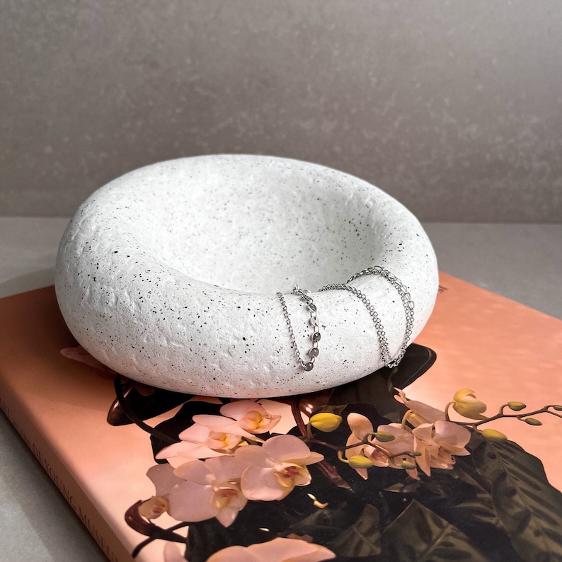 May include: White speckled ceramic bowl with a rounded, organic shape. Two silver chains are resting inside the bowl.