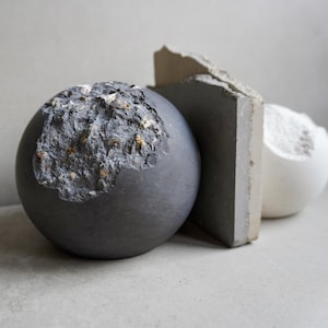 Puede incluir: Una composici&oacute;n de naturaleza muerta con tres esculturas abstractas. Una esfera gris oscuro con una superficie texturizada y erosionada se coloca junto a una losa rectangular gris y una forma esf&eacute;rica blanca. Los objetos se presentan sobre un fondo neutro.