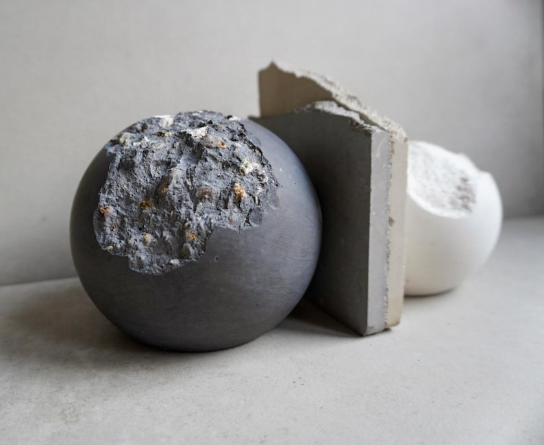 Puede incluir: Una composici&oacute;n de naturaleza muerta con tres esculturas abstractas. Una esfera gris oscuro con una superficie texturizada y erosionada se coloca junto a una losa rectangular gris y una forma esf&eacute;rica blanca. Los objetos se presentan sobre un fondo neutro.