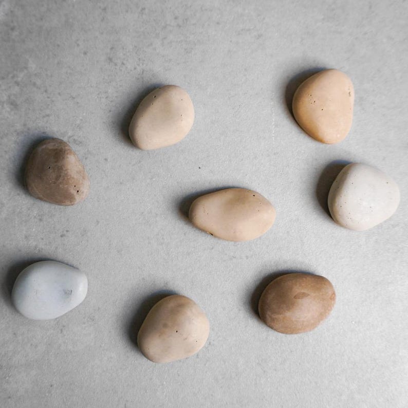 K&ouml;nnte beinhalten: Draufsicht auf neun glatte, ovale Steine in verschiedenen Beige-, Braun- und Wei&szlig;t&ouml;nen. Die Steine sind auf einer hellgrauen Oberfl&auml;che angeordnet und erzeugen eine nat&uuml;rliche, minimalistische &Auml;sthetik. Die Steine variieren in Gr&ouml;&szlig;e und Farbe.