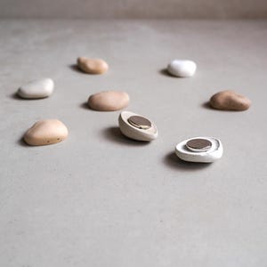 K&ouml;nnte beinhalten: Eine Sammlung von steinartigen Magneten in verschiedenen neutralen Farben, darunter Beige, Wei&szlig; und Braun. Zwei Magnete sind aufgespalten und zeigen eine silberne Scheibe. Die Magnete sind auf einer hellgrauen Oberfl&auml;che angeordnet.