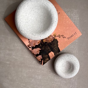 May include: Two white speckled ceramic bowls, one larger than the other, on a pink and white book with the text "Bouquets" and "Pamela J".